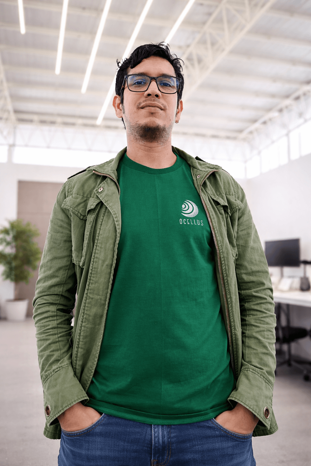 Hombre con gafas, camiseta verde de Ocellus y chaqueta, posando en una oficina moderna.